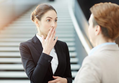 Young employee keeping her palm on mouth and expressing amazement while talking to her colleague